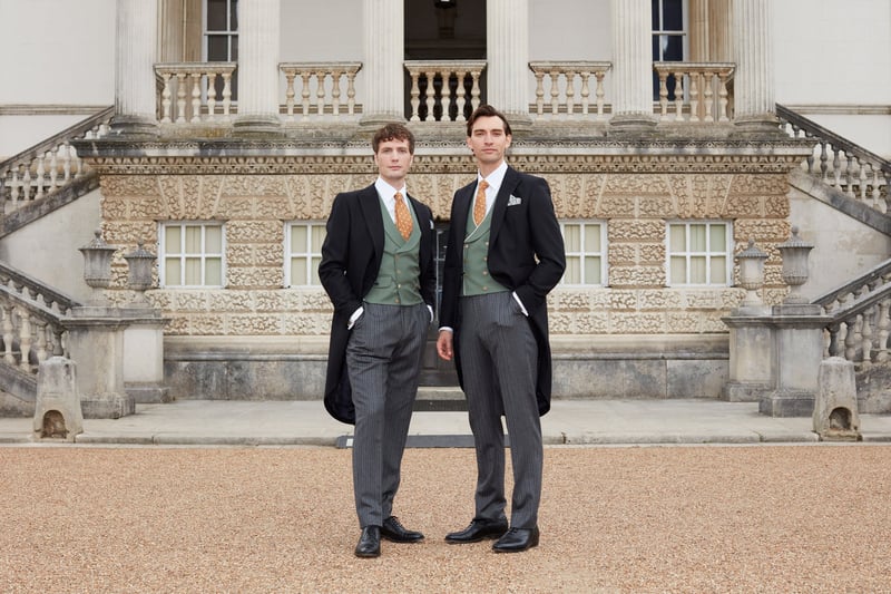 two male models wearing traditional morning suits with green waistcoats stand in front of a grand venue.