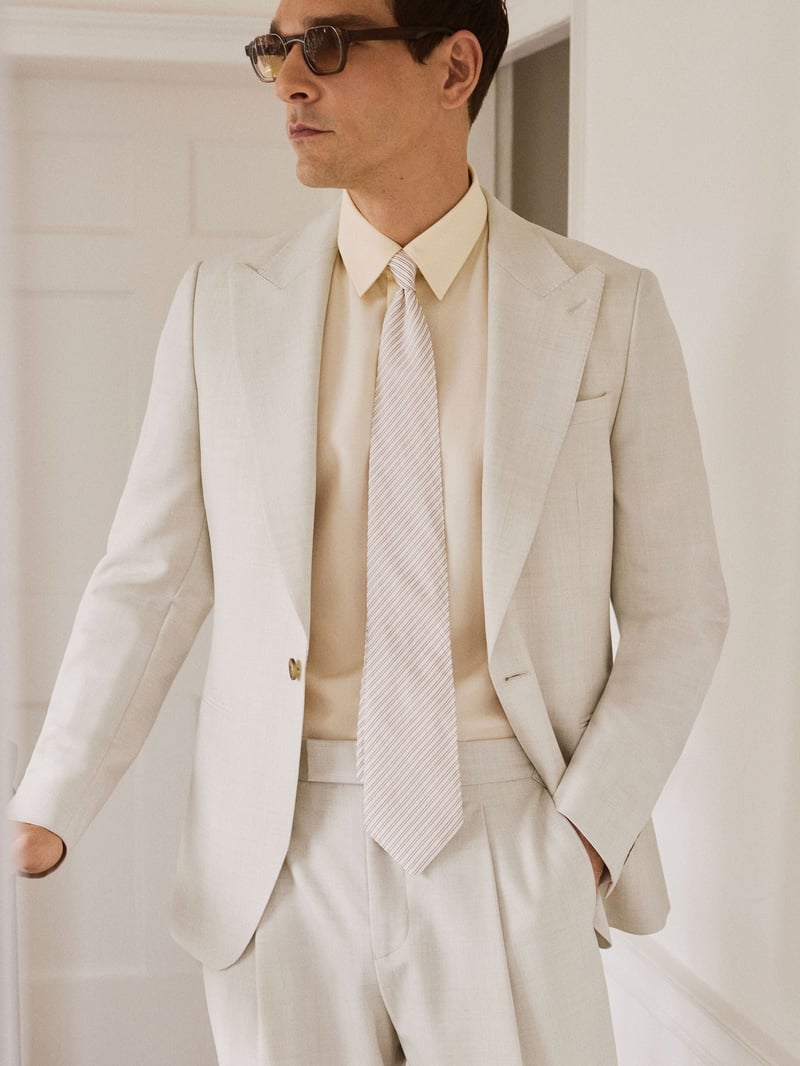 a male model wearing a stone coloured wedding suit in linen with a matching tie and sunglasses.