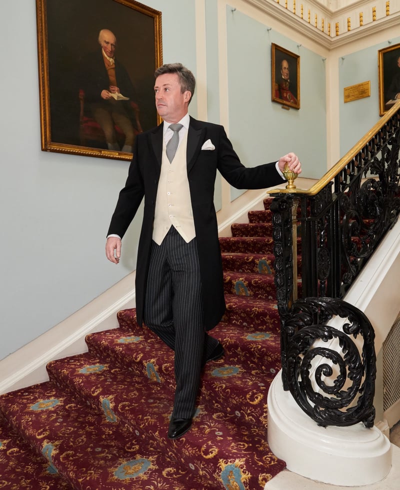 a man in a black morning suit and cream waistcoat walks down the stairs of a stately home