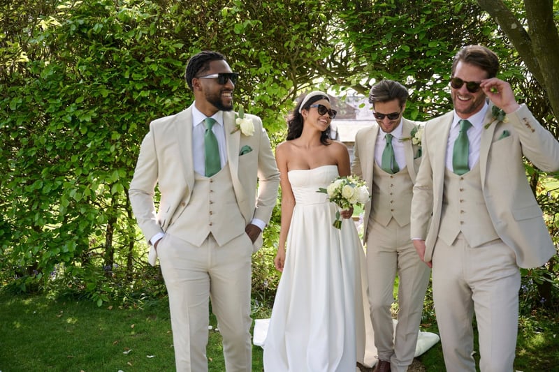 a bride and three groomsmen in cream-coloured suits with green ties all smiling and wearing sunglasses at a summer wedding.
