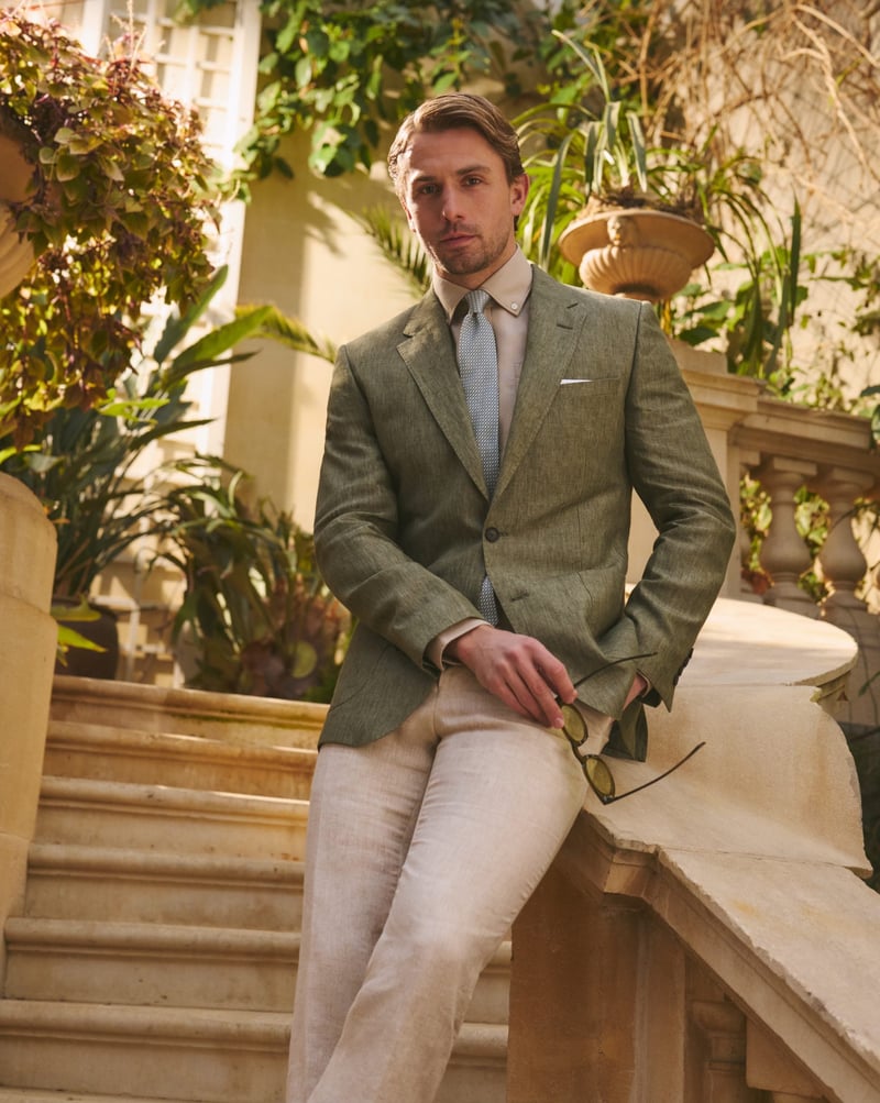 a male model in a tropical destination wearing an olive green summer suit jacket with cream trousers and light blue tie.