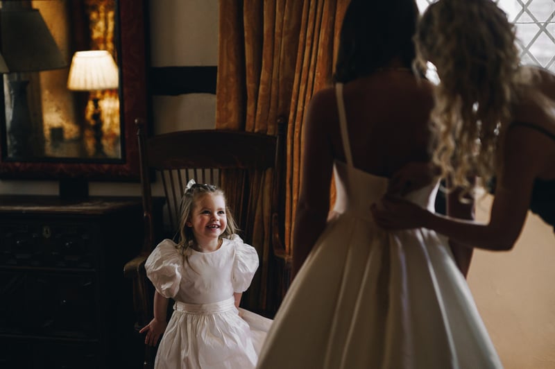 a flower girl in a white dress smiles as the bride gets ready.