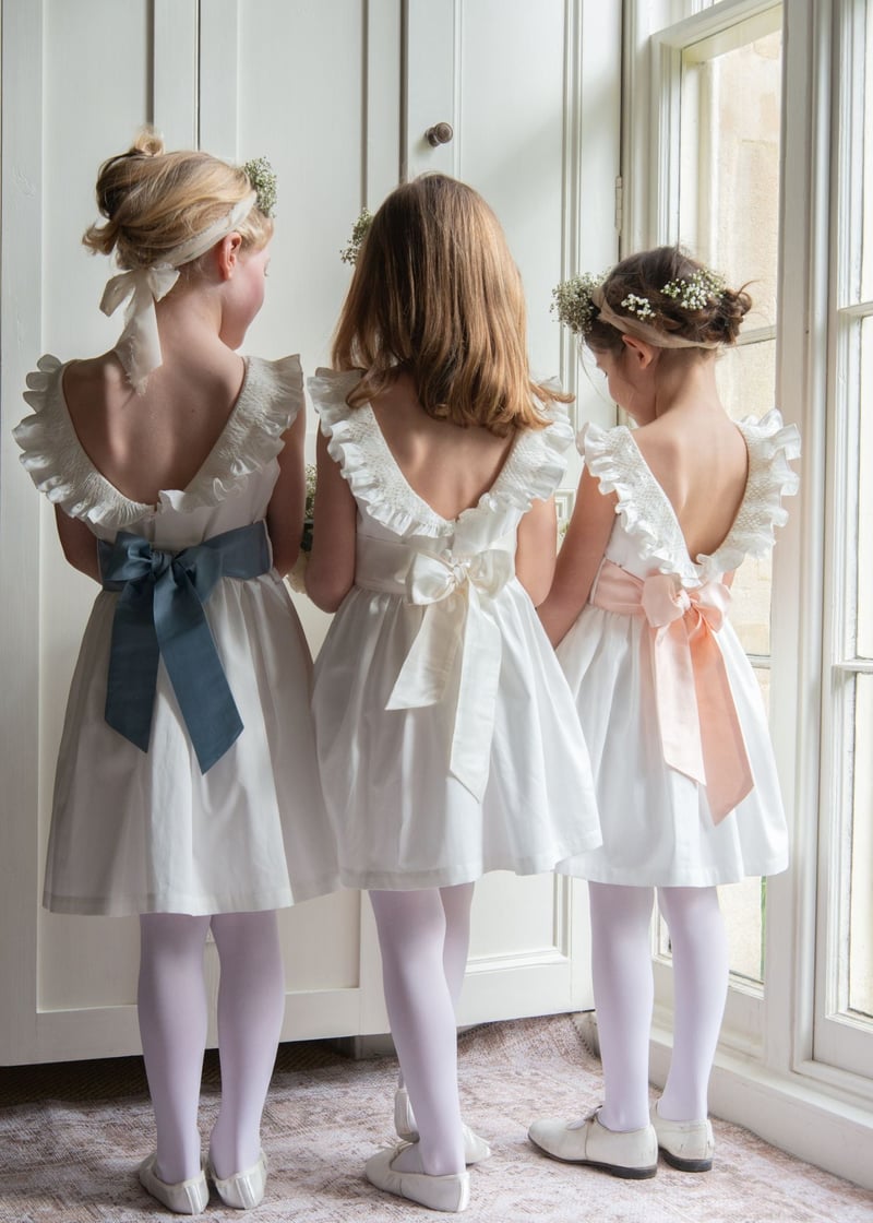 three flower girls in white smocked dresses and different coloured sashes stand with their back to the camera. 