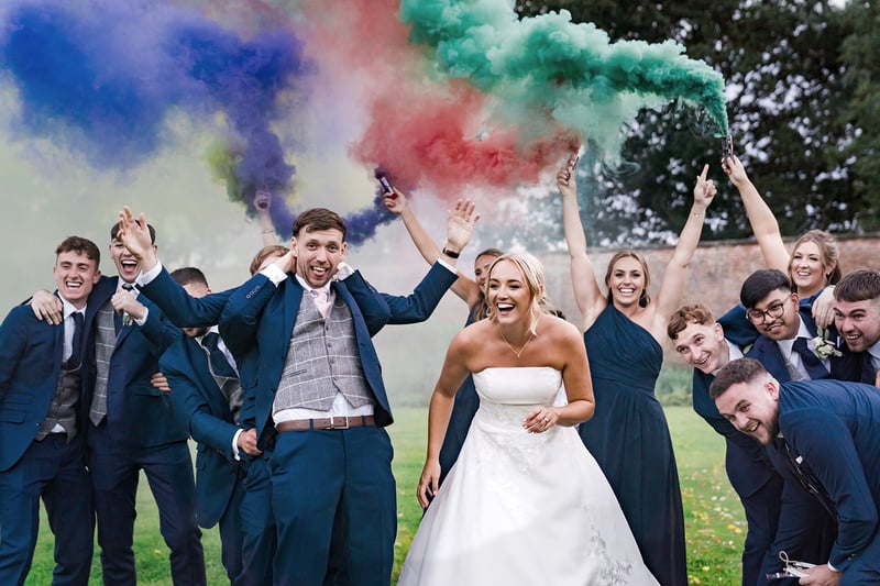 a bride and groom smile as their wedding party hold up colourful flares.