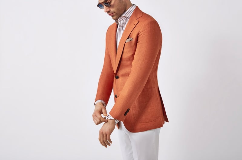 a male model wears an orange tailored jacket with white trousers, striped shirt and sunglasses.