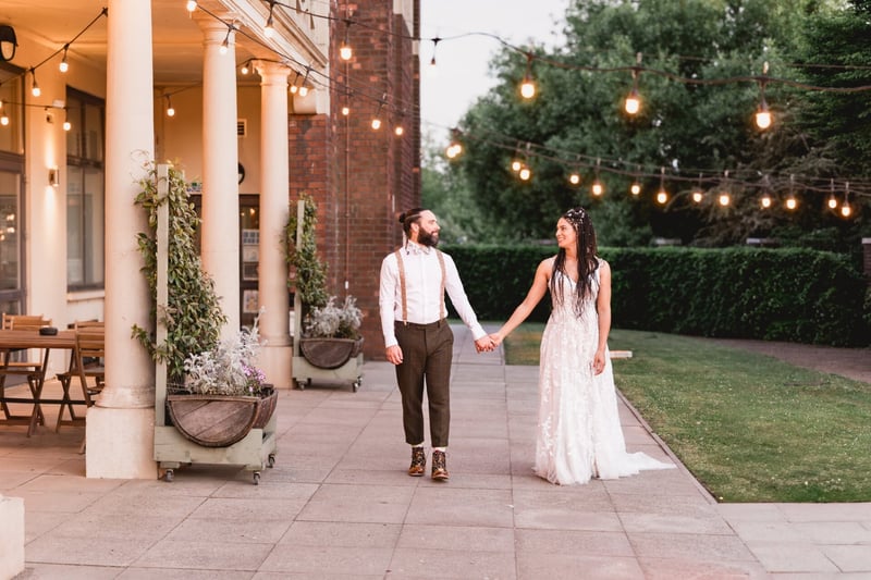 a newlywed bride and groom look at each other and smile as they hold hands in an outdoor setting with festoon lights.