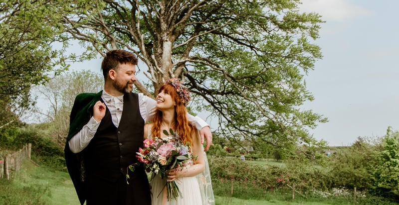 a bride holding colourful flowers and wearing a flower crown walking with her groom from their humanist ceremony.