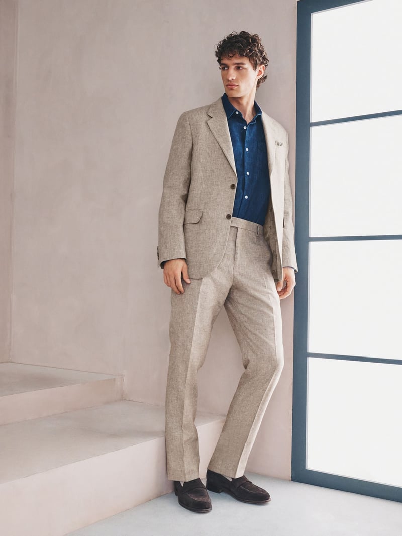 a male model leans against a window wearing a natural-coloured linen suit with dark blue shirt.
