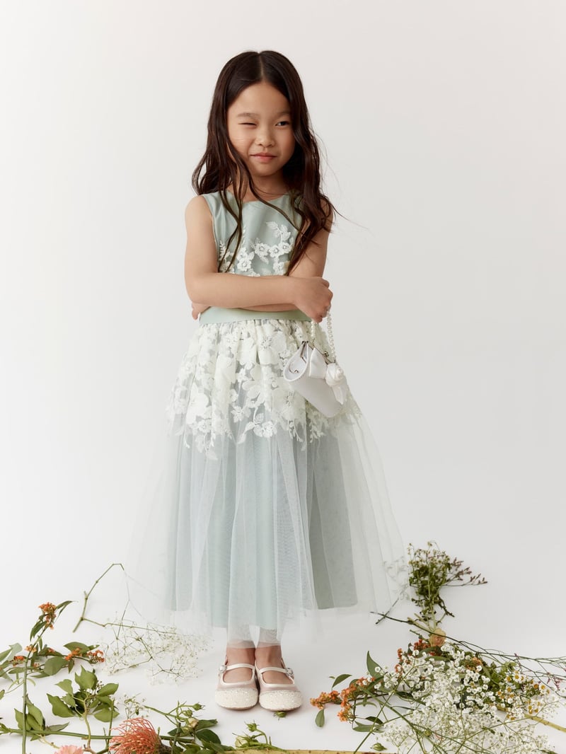a child model wears a sage green flower girl dress with white lace detail.