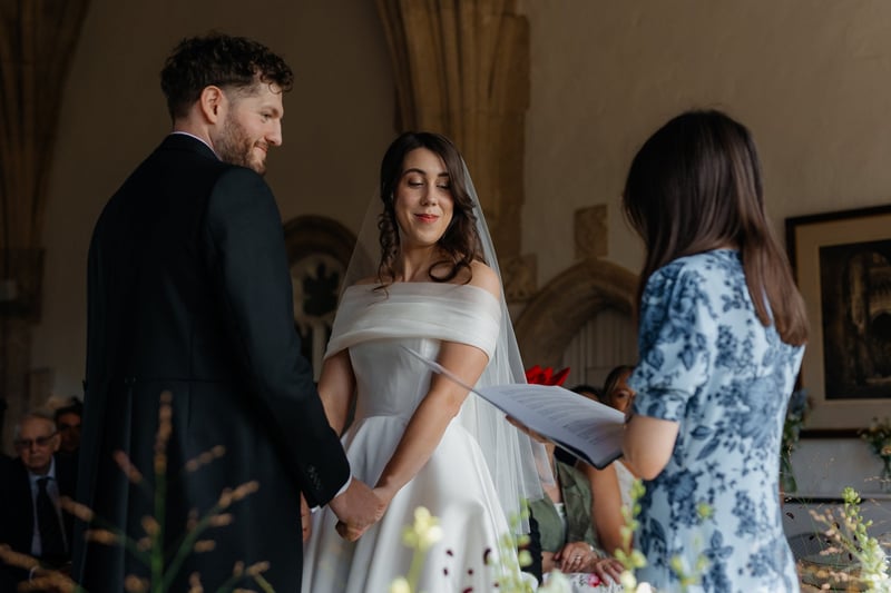 a bride smiles as she holds hands with her groom at the altar and looks down at the words the registrar is reading from a sheet of paper.