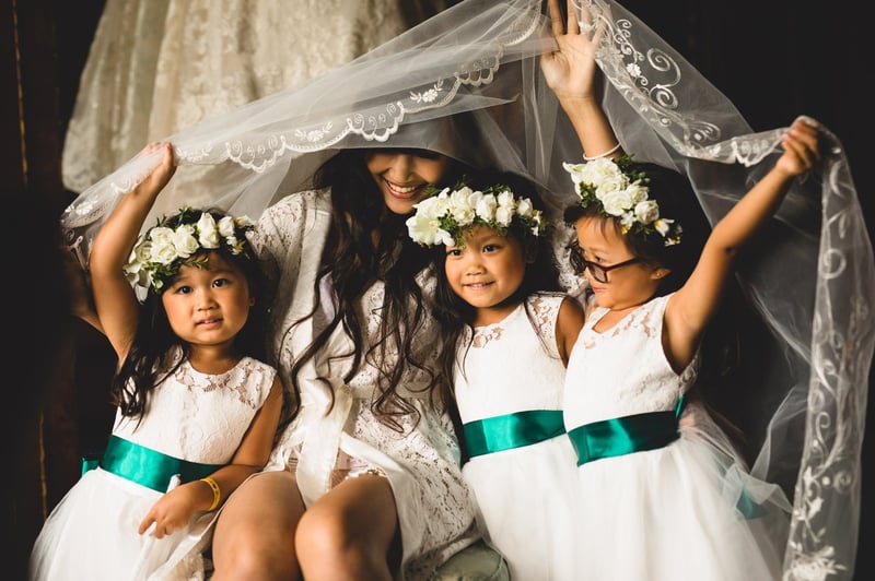 a bride and three flower girls in flower crowns smile as they hold a wedding veil.