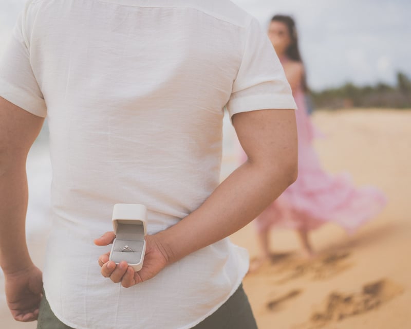 a couple stand on a beach and in the foreground a man holds an engagement ring in a ring box behind his back.