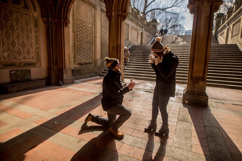 a man in a coat and hat on bended knee as his proposes to his partner in a city. Both are wearing winter clothes.