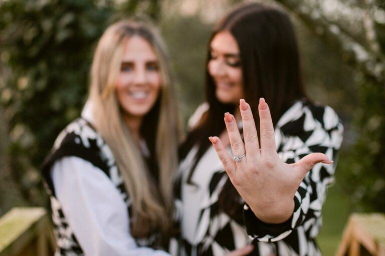 A person extends their left hand toward the camera to show an engagement ring, with a same sex couple standing blurred in the background outdoors