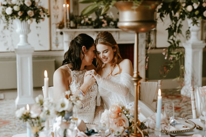 two brides with half-up half-down hairstyles sitting at a table dressed for a wedding breakfast.
