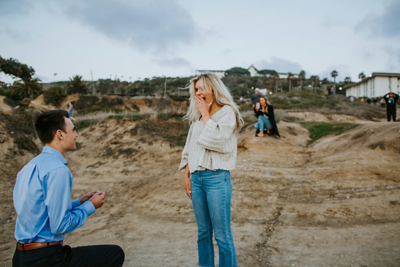 a man down on one knee proposing to a woman. in the background a woman captures it on camera.