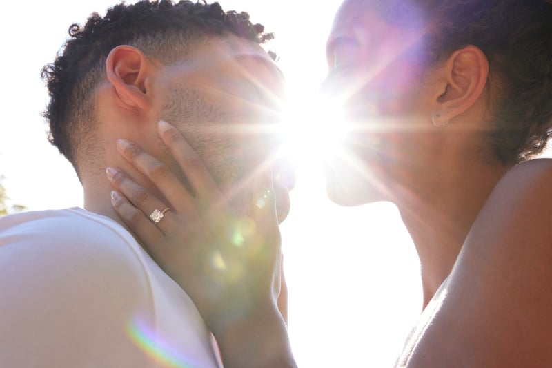a woman wearing an engagement ring holding her partner's face in her hands as the sun shines in the background.
