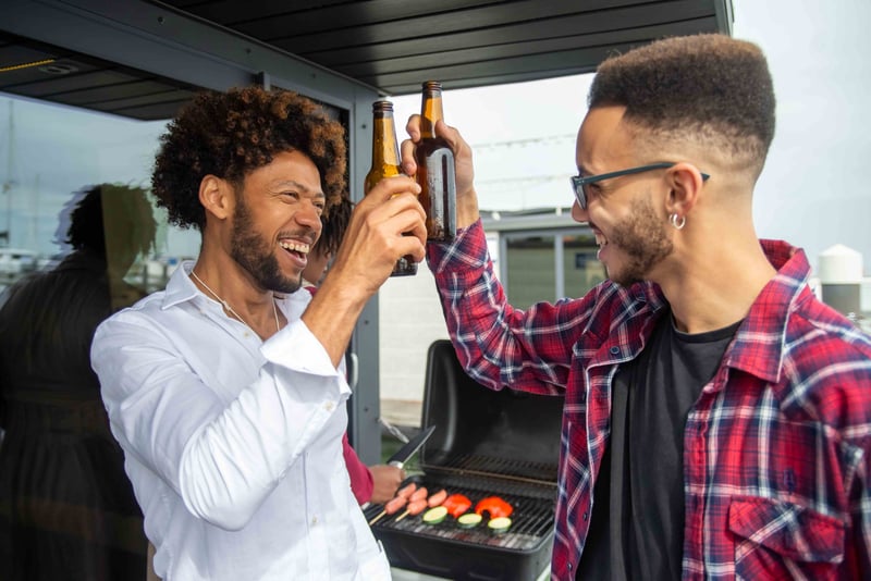 two men clinking bottles and laughing as they stand next to a barbecue.
