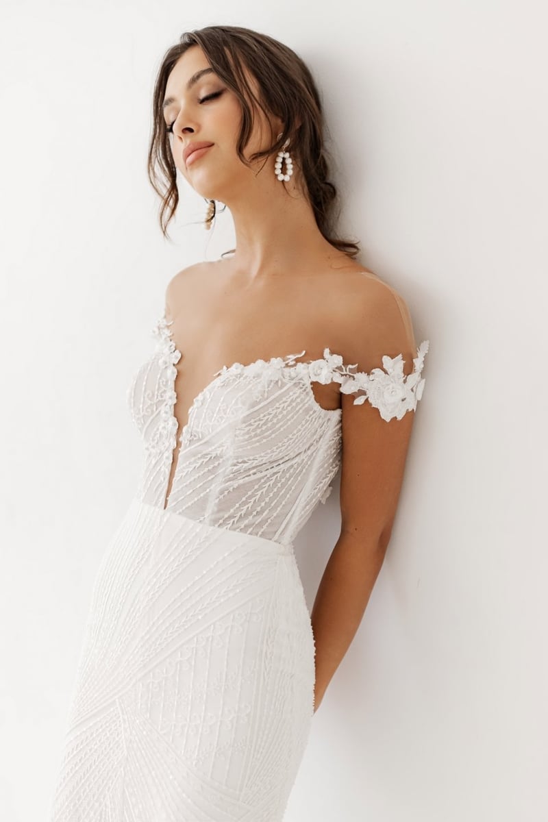 a model wears an off the shoulder wedding dress with sheer panel corset detail and plunging neckline.