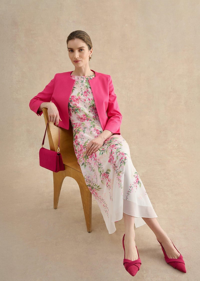 a model sitting down wears a floral midi dress with lightweight pink tailored jacket.