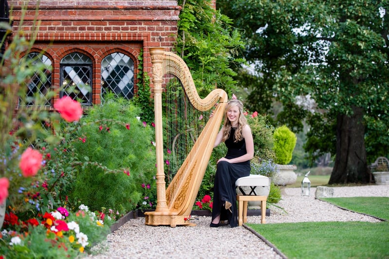 a harpist sits in a garden wedding venue smiling at the camera next to her harp.