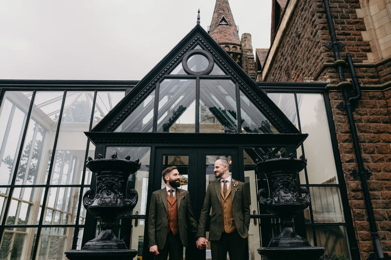 two grooms holding hands in front of their hotel wedding venue.