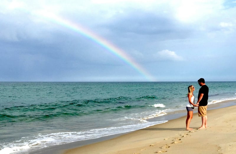 a man and a woman looking at each other and holding hands on a beach. in the background is a rainbow.