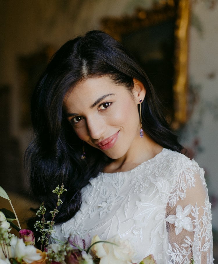 a bride with dark hair and a fabulous blow-dry looks into the camera as she holds her wedding bouquet.