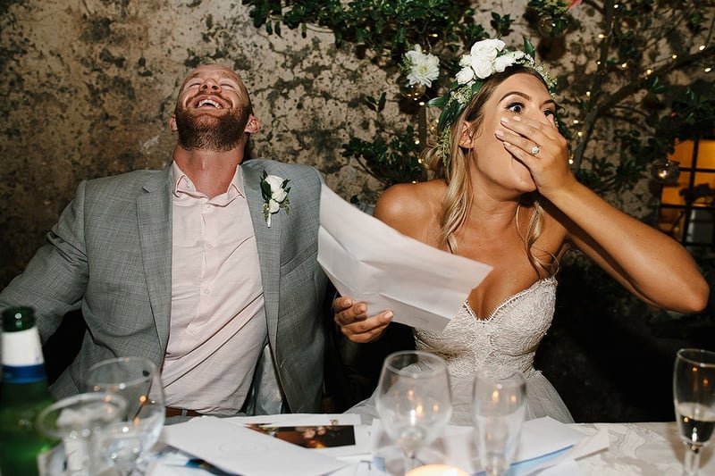 a bride holding a piece of paper with her hand over her mouth in shock as the groom next to her laughs.