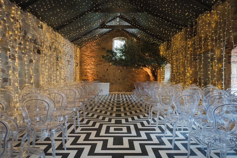 interior of a stone barn like room with jazzy black and white floor, fairy lights and clear perspex chairs set up for a wedding ceremony.