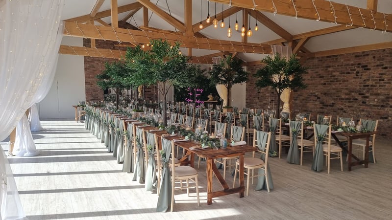 interior of a modern barn wedding venue with long tables, hanging lights and tall green tree tablecentres.