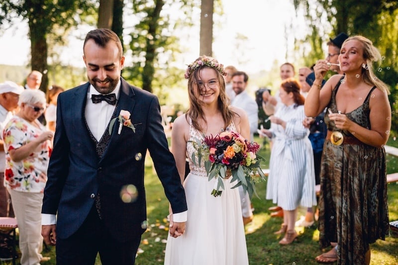 Glückliches Hochzeitspaar im Wald umgeben von Hochzeitsgästen, die Seifenblasen machen
