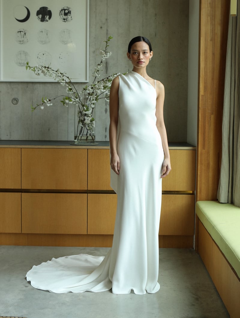 a model standing in a room with artwork on the wall wears a one-shoulder silk crepe wedding dress.