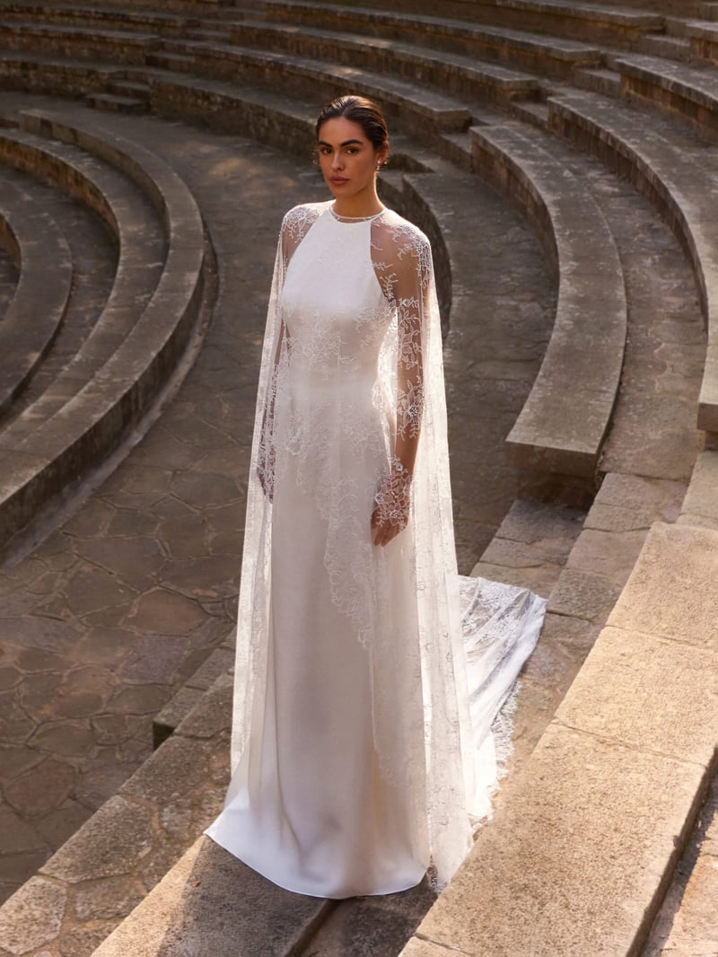 a model standing on stone steps wearing a halter wedding gown with open back and detachable cape