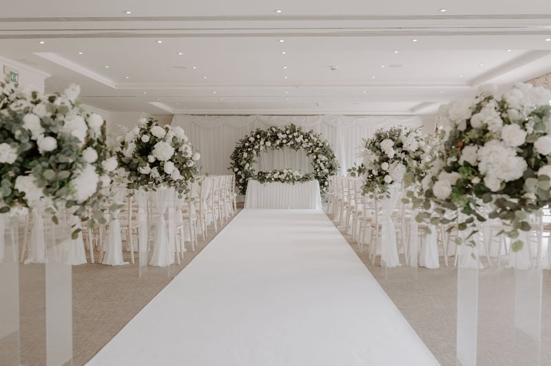 interior of a country house hotel function room with white decor and green and white floral installations set up for a ceremony.