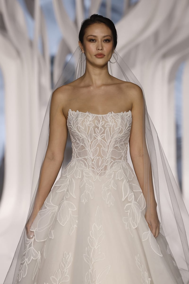 a model on a runway wears a strapless embroidered tulle ballgown wedding dress with embroidered soutache tulle appliqués.