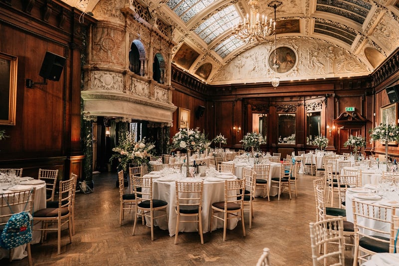 a wedding breakfast set up in a grand hall of a historic wedding venue, with high arched ceilings and round tables with tall centrepieces.