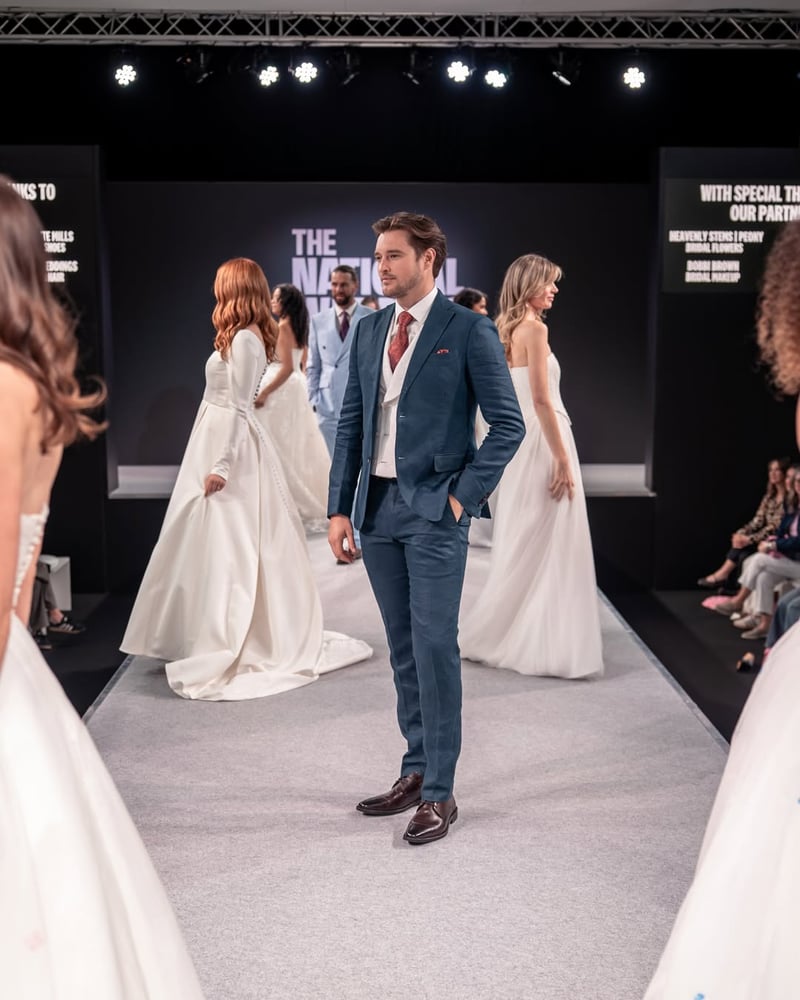 a male model in a suit surrounded by female models in wedding dresses on the catwalk at the national wedding show.