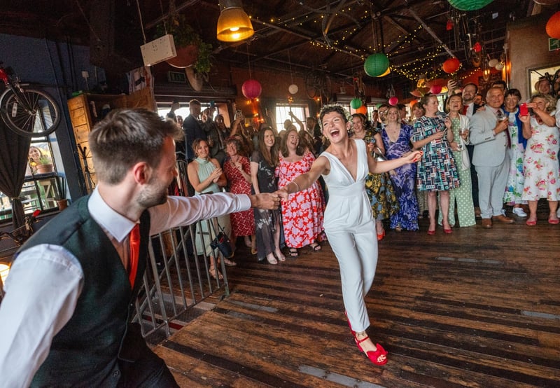 a happy bride wearing a white trouser suit dancing to an upbeat wedding first dance song with her groom as guests watch and clap. There are colourful lanterns on the ceiling.