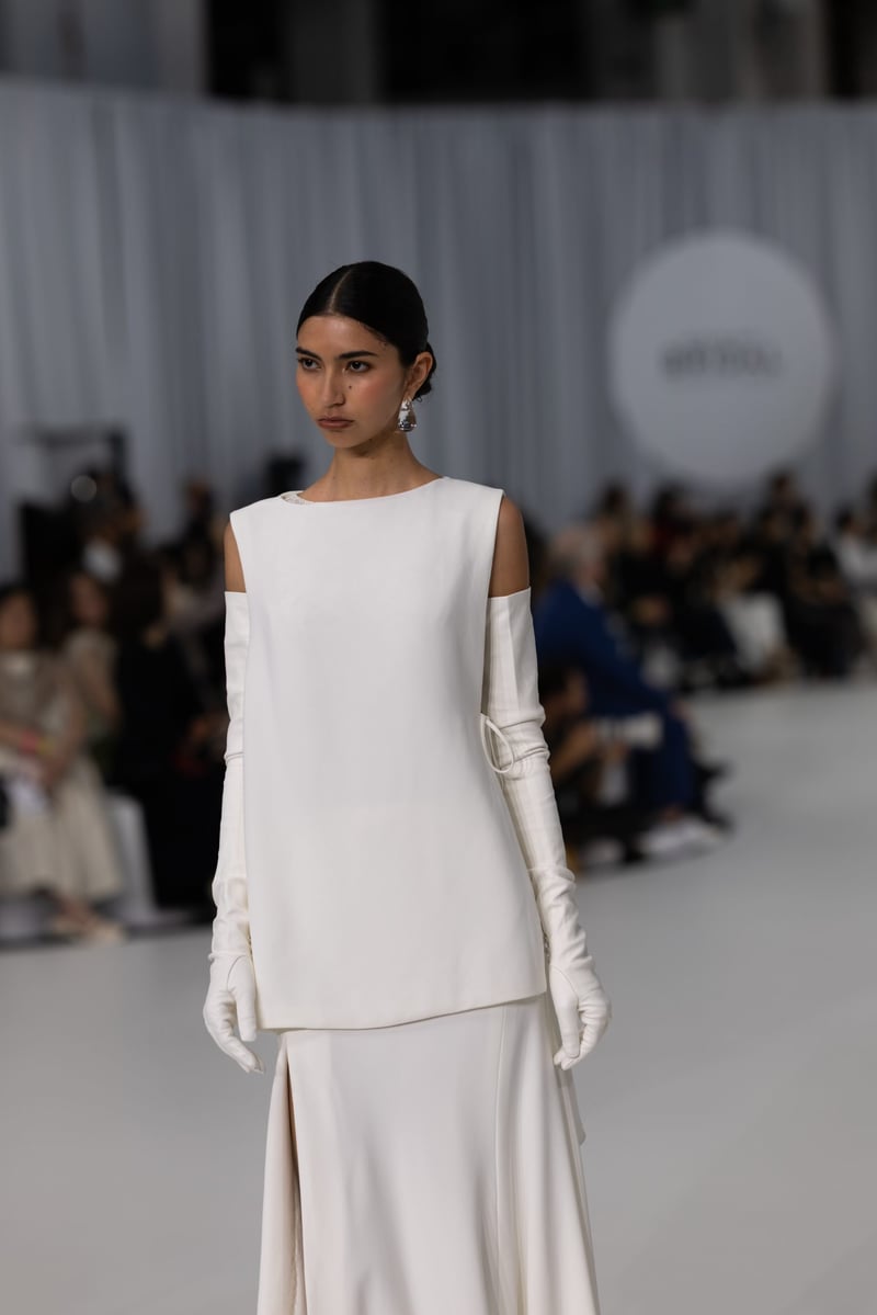 a model on the runway wears a minimal straight-neck bridal gown crafted in fluid crepe with long sleeve crepe gloves.