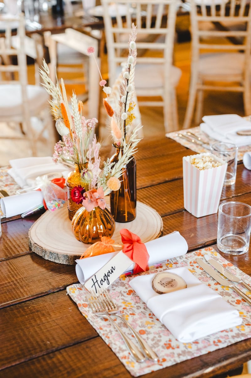 a long slice featuring a bud vase arrangement of dried flowers on a wooden table with ditsy floral print place mat and wood slice name tag.