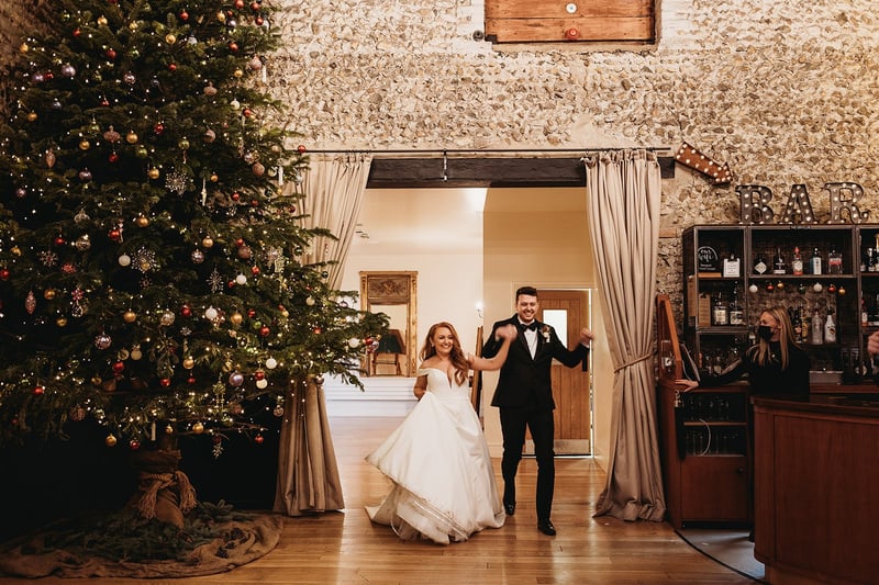 a bride and groom walk into a barn wedding venue hand in hand next to a giant Christmas tree.