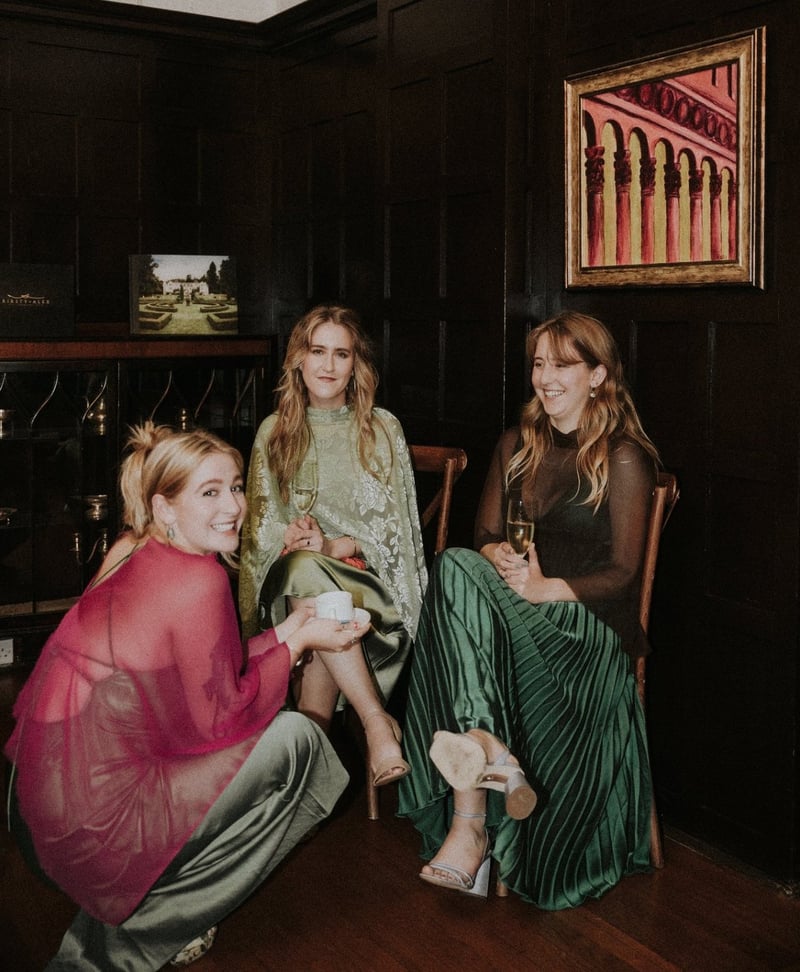 three female wedding guests in winter wedding guest dresses huddle together with their drinks and smile in a polaroid style snapshot.