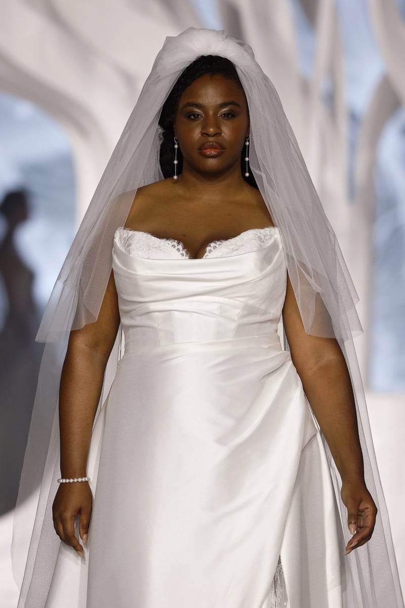 a plus size model walks along a runway wearing a stretch mikado a-line Plus Size gown with draped strapless neckline and ruched skirt.