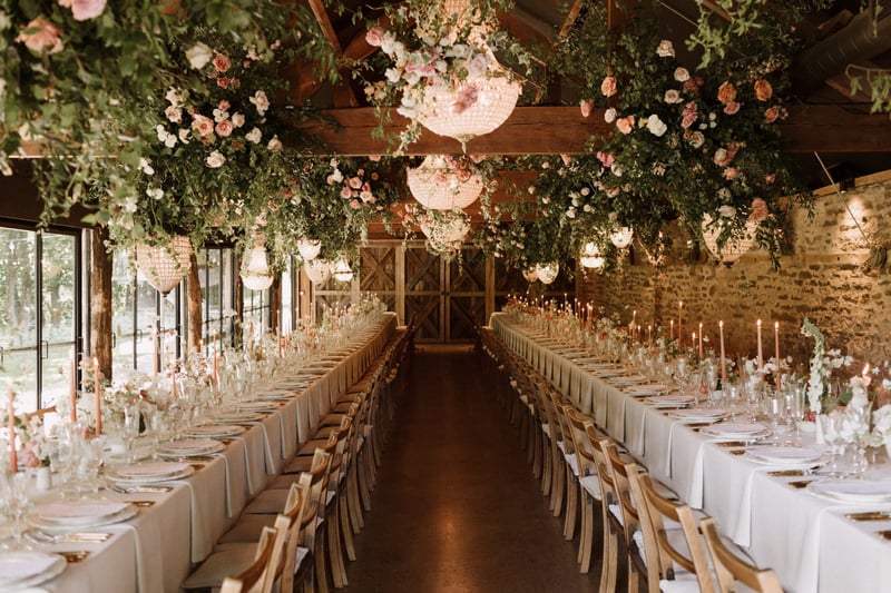 interior of a stone barn wedding venue with tables set up for a wedding breakfast and floral installations on the ceiling. 