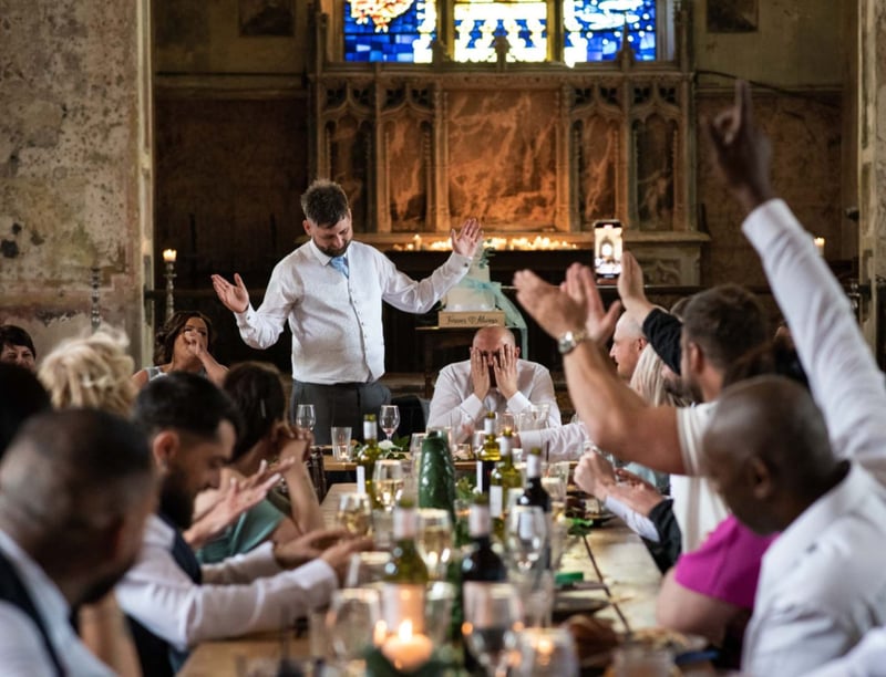 a groom at the end of a long table holds his face in his hands as his best man stands and holds his arms in the air during his speech.