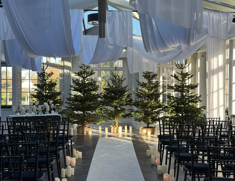 a ceremony set up for a Christmas wedding with black chairs, white drapes and a line of Christmas trees and pillar candles.