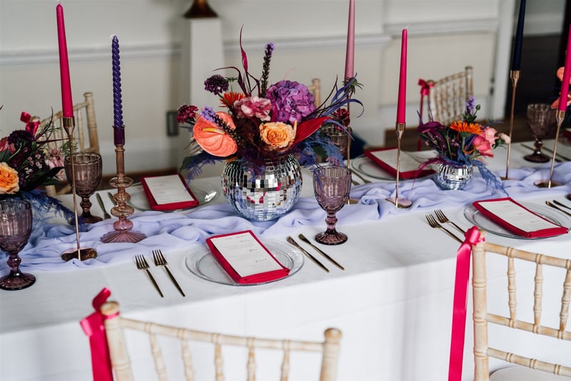 a modern wedding table set-up with pink and purple taper candles, disco balls filled with bright flowers, purple glassware, and pink and white wedding menus.