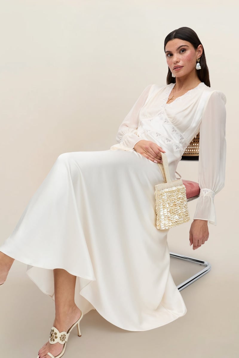 a model sitting back in a chair wears a vintage-style silk-satin midi wedding dress with long sleeves.