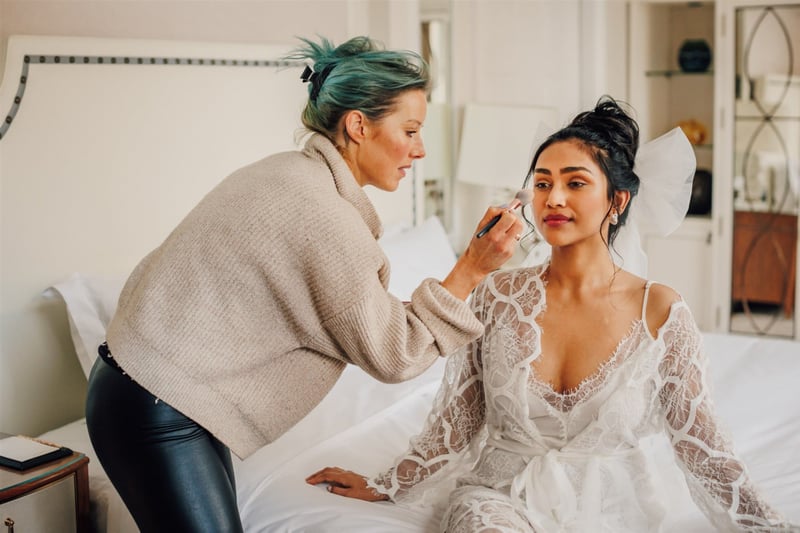 a makeup artist applies a bride's wedding makeup as she sits on a bed.
